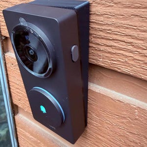 May include: A black rectangular doorbell camera mounted on a brown wooden surface. The camera has a circular lens and a button on the side. A blue light is visible on the bottom of the device.