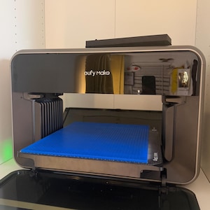 May include: A eufy Make 3D printer with a metallic bronze frame and a glossy black front panel. The device features an open build chamber with a bright blue square print bed surface. A horizontal glass door is positioned at the bottom of the unit.