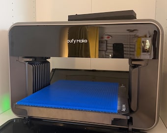 May include: A eufy Make 3D printer with a metallic bronze frame and a glossy black front panel. The device features an open build chamber with a bright blue square print bed surface. A horizontal glass door is positioned at the bottom of the unit.