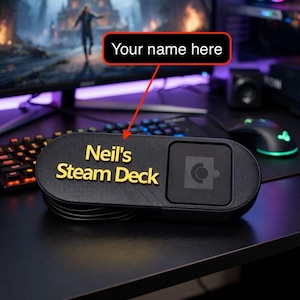 May include: Black Steam Deck case with the text "Neil's Steam Deck" in gold lettering. The case has a rounded rectangular shape with a square section on the right side. A keyboard, mouse, and monitor are visible in the background.