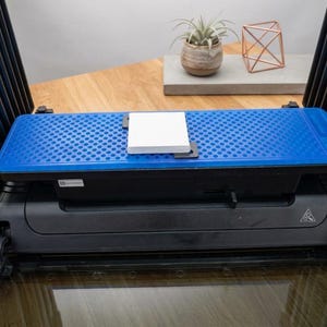 May include: A black electronic device featuring a rectangular blue top surface with a honeycomb pattern. A small, square white plate is centered on the blue surface. The device has black vertical side panels and a black base with a warning symbol.
