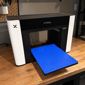 May include: A black and white xTool laser machine featuring a prominent blue honeycomb work bed extending from the front. The device has a sleek, rectangular frame with the brand name XTOOL printed in white on the front panel and a green X logo.