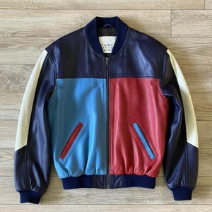 Puede incluir: Chaqueta bomber de cuero con bloques de color en azul marino, azul claro y rojo. La chaqueta tiene mangas blancas y cuello y puños azul marino. Cuenta con dos bolsillos delanteros con detalles en rojo y azul.