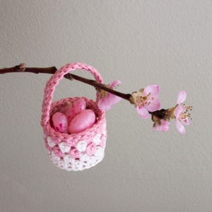 A small crochet basket in pink and white is hanging on a peach branch. The basket is filled with small candy of the same colour.