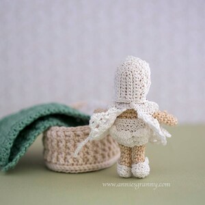 Crochet baby doll with white dress and cap, seen from behind. Basket with a blanket and a pillow.
