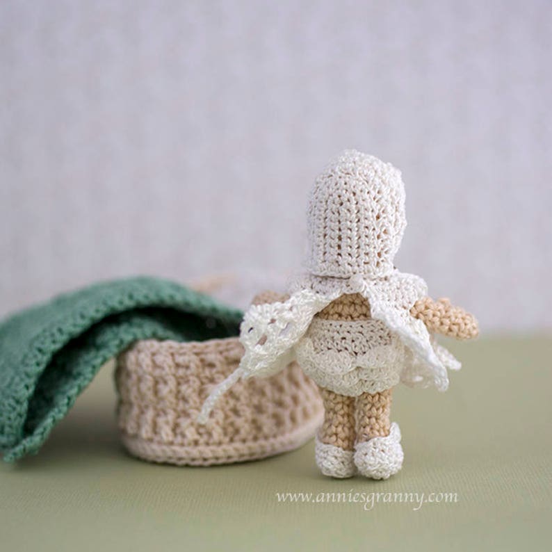 Crochet baby doll with white dress and cap, seen from behind. Basket with a blanket and a pillow.
