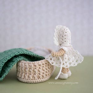 Crochet baby doll with white dress and cap, seen from the side. Basket with a blanket and a pillow.