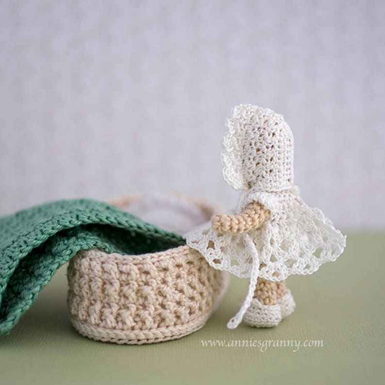 Crochet baby doll with white dress and cap, seen from the side. Basket with a blanket and a pillow.
