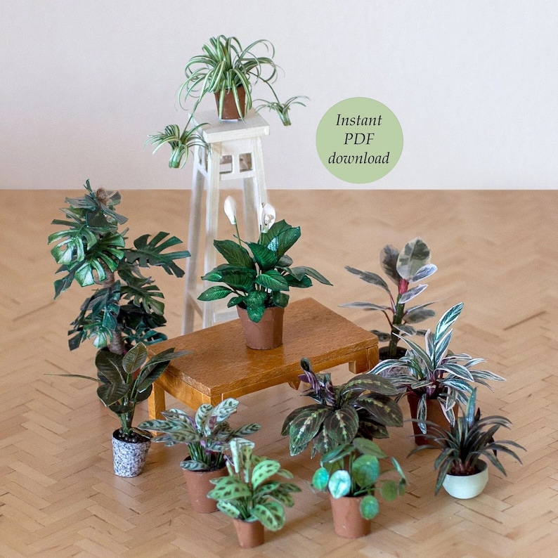 Ten miniature paper plants for dollhouses.  A spider plant is placed on a white pedestal. A peace lily is placed on a brown table in front of the pedestal. The other plants are on a wooden floor.