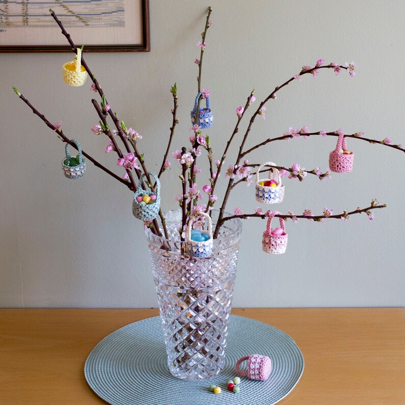 The picture shows a large vase with peach twigs. On the twigs hang small crocheted baskets filled with sweets. A basket has fallen on the side of the vase and the candy has fallen out.