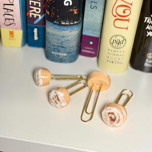 Könnte beinhalten: Vier Zimtschnecken-förmige Büroklammern mit goldfarbenen Metallclips. Die Miniaturbrötchen haben eine hellbraune Basis mit einer weißen Zuckerguss-Wirbel. Die Clips sind auf einer weißen Oberfläche in der Nähe einer Reihe bunter Bücher angeordnet.