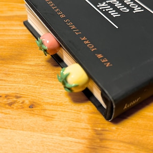 May include: A black book with the title "milk and honey" and "NEW YORK TIMES BESTSELLER" text. Two tomato-shaped bookmarks, one pink and one yellow, are inserted into the book. The book is on a wooden surface.