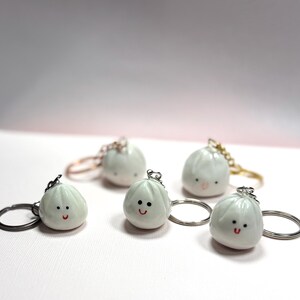 May include: A collection of white dumpling-shaped keychains with smiling faces. Each keychain has a silver or gold-colored ring. The dumplings have small black dots for eyes and a red curved line for a mouth.