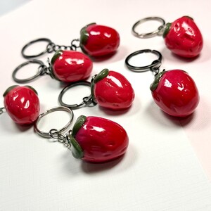 May include: A selection of red strawberry keyrings. Each keyring features a glossy, vibrant red strawberry with a green stem and a silver-coloured keyring. The keyrings are scattered on a white and pink surface.
