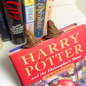 May include: Two brown Sorting Hat bookends sit atop a red "Harry Potter and the Philosopher's Stone" book. The bookends are detailed with a hat shape. Other books are visible in the background.