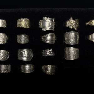 May include: A collection of silver rings with various designs, including floral patterns, architectural elements, and abstract shapes. The rings are displayed on a black surface, showcasing their intricate details and textures.