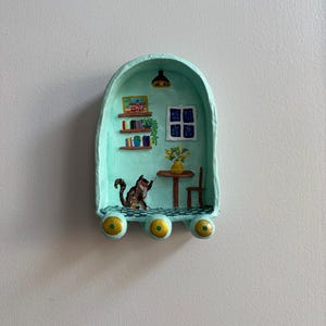 May include: A miniature, hand-painted diorama in a teal arch shape. Inside, a cat reaches toward a table with a vase of flowers. A bookshelf, window, and hanging light complete the cozy scene. The base has three gold and teal spheres.