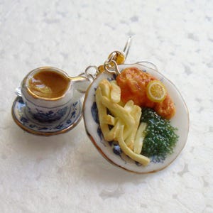 Puede incluir: Charm en miniatura que representa una taza de té y un plato de fish and chips. La taza y el platillo tienen un diseño floral azul y blanco. El plato incluye pescado frito, patatas fritas, una rodaja de limón y verduras verdes.