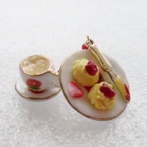 May include: A miniature charm featuring a white teacup with a strawberry design on a matching saucer, paired with a white plate holding two yellow scones topped with red jam, a slice of strawberry, and a small gold-toned butter knife.