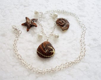 Puede incluir: Un collar de cadena plateada con dijes de conchas marrones y blancas, incluyendo una estrella de mar, una almeja y una concha de nautilus. El collar también presenta acentos de perlas blancas.