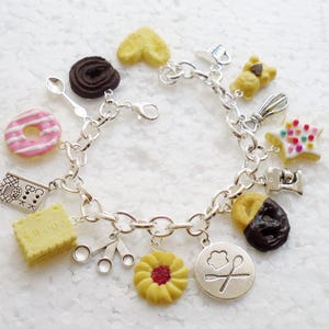 May include: A silver charm bracelet with various food-themed charms, including a donut, a cookie, a heart, a star, and a spoon. The charms are attached to a silver chain bracelet.