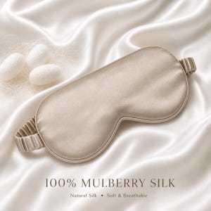 May include: A beige silk eye mask with an elastic band, resting on a satin fabric. The text "100% MULBERRY SILK" is displayed below the mask, along with "Natural Silk &bull; Soft & Breathable". Three white silk cocoons are in the upper left corner.