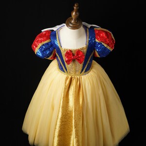 May include: A dress featuring a blue sequined bodice with gold trim, red sequined puff sleeves, and a red bow at the center neckline. The skirt consists of a gold fabric panel layered over a full, voluminous yellow tulle skirt with a white collar.