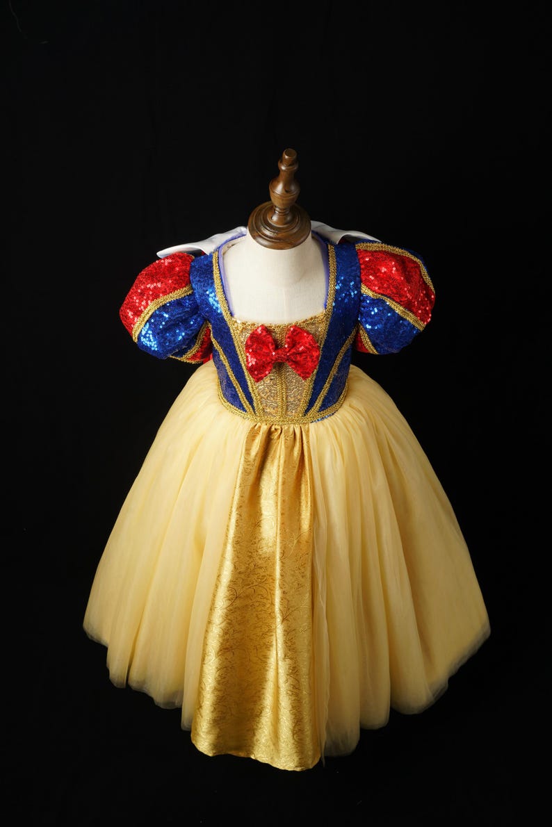 May include: A dress featuring a blue sequined bodice with gold trim, red sequined puff sleeves, and a red bow at the center neckline. The skirt consists of a gold fabric panel layered over a full, voluminous yellow tulle skirt with a white collar.