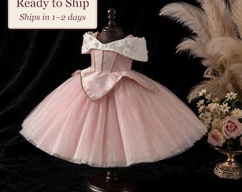Op de afbeelding: Een roze jurk met een off-shoulder witte kraag versierd met kleine witte bloemapplicaties, een gestructureerd lijfje met verticale naden en een volumineuze tule rok met een subtiel wervelend patroon op een donkere standaard.