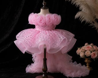 Op de afbeelding: Een lichtroze jurk met een lijfje versierd met grote organza rozetten langs de halslijn, een middengedeelte met lovertjes en een volumineuze high-low rok met meerdere lagen ruches van tule die tot op de grond vallen.