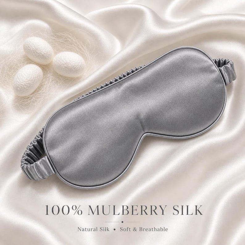 May include: A gray silk sleep mask with an elastic band, resting on a white satin surface. The text "100% MULBERRY SILK" is displayed below the mask, with the words "Natural Silk &bull; Soft & Breathable".