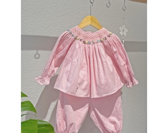 Op de afbeelding: Een lichtroze set bestaande uit een top met lange mouwen en een bijpassende broek. De top heeft een gesmokte halslijn met fijn bloemenborduursel in blauw, geel en groen. De mouwen en broekspijpen zijn afgewerkt met geplooide boorden.