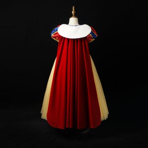 May include: A floor-length dress featuring a long red velvet cape with a white collar, puffed sleeves with blue and red sequins, and a full-length yellow tulle skirt. The garment is displayed on a mannequin against a solid black background.