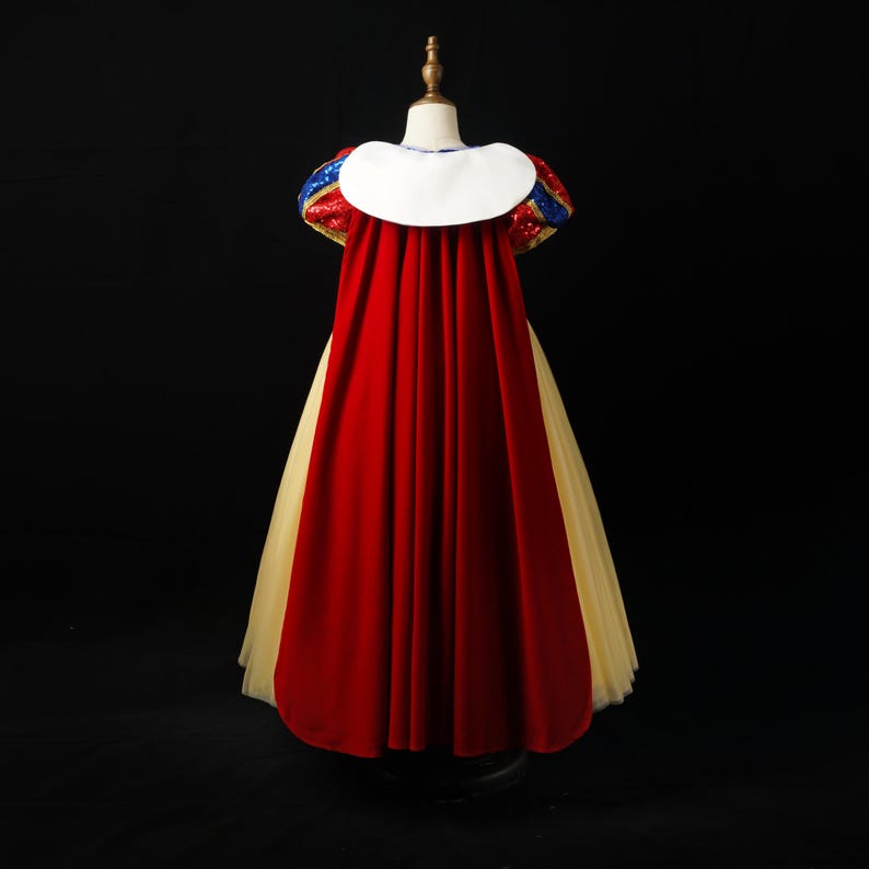 May include: A floor-length dress featuring a long red velvet cape with a white collar, puffed sleeves with blue and red sequins, and a full-length yellow tulle skirt. The garment is displayed on a mannequin against a solid black background.