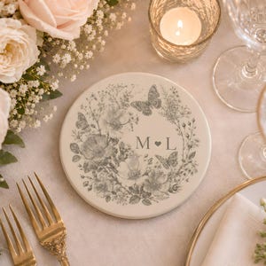 May include: A round, off-white ceramic coaster featuring a grey botanical illustration of wildflowers and butterflies arranged in a circular wreath. The center of the design displays the letters M and L separated by a small heart symbol.