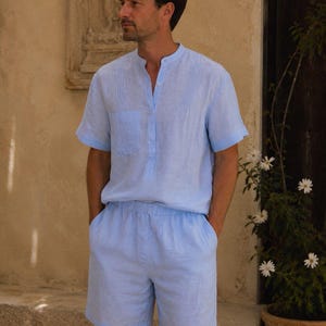May include: A light blue short-sleeve shirt with a mandarin collar, a front button placket, and a chest pocket, paired with matching light blue shorts featuring an elastic waistband. The set is made of a textured fabric with vertical pleat details.