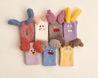 Op de afbeelding: Een collectie van zeven pluizige, pluche sokken ontworpen als dierenfiguren. Elke sok heeft unieke kleuren, waaronder roze, wit, bruin, paars, lavendel, blauw en geel, met geborduurde gezichtsdetails en bevestigde 3D-oren.