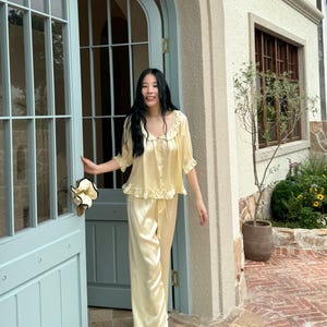 May include: A pale yellow two-piece pajama set consisting of a button-down top with a ruffled collar and short sleeves, paired with matching wide-leg trousers featuring a drawstring waist and ruffled hems, crafted from a smooth, satin-like fabric.
