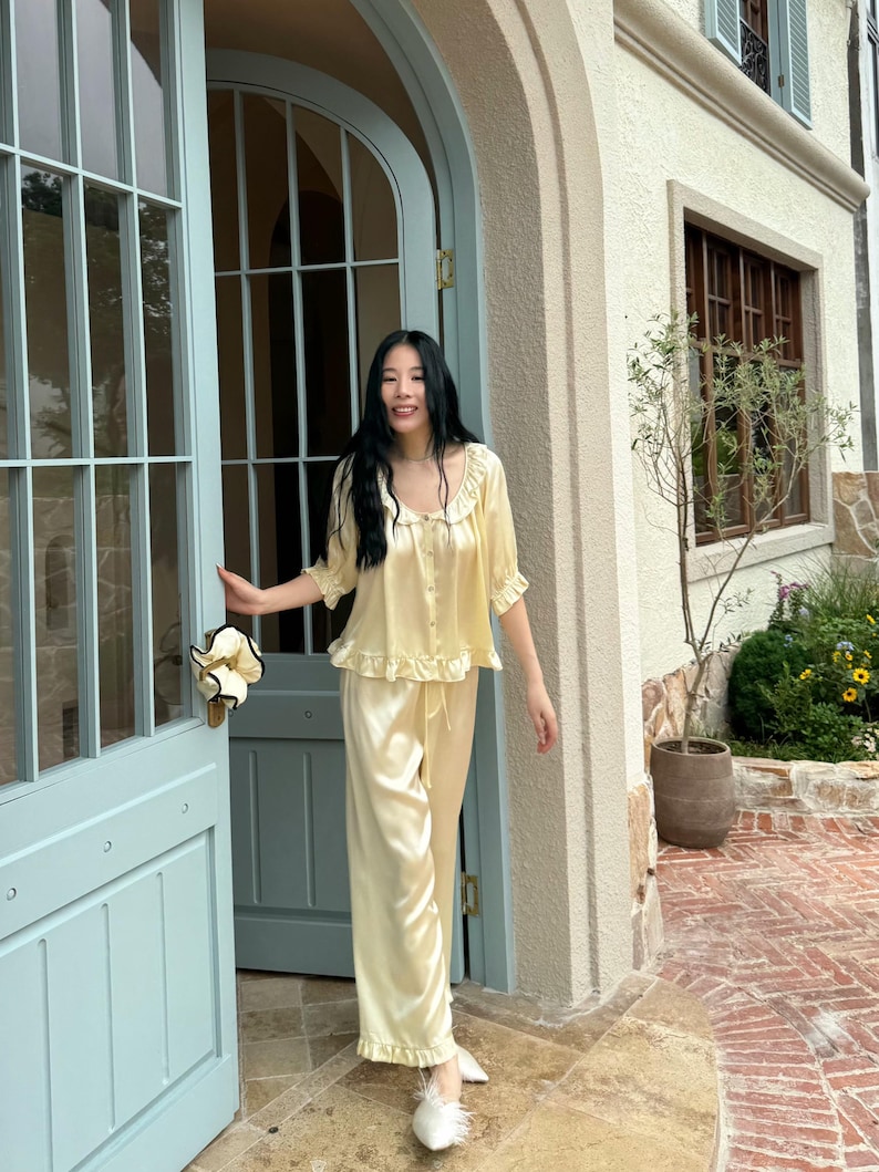 May include: A pale yellow two-piece pajama set consisting of a button-down top with a ruffled collar and short sleeves, paired with matching wide-leg trousers featuring a drawstring waist and ruffled hems, crafted from a smooth, satin-like fabric.
