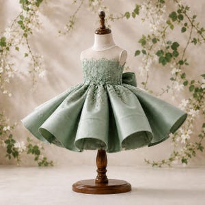 May include: A sage green dress featuring a sleeveless bodice with intricate lace embroidery and floral appliqués. The skirt is voluminous and pleated, extending from the waist. A large bow is attached to the back of the dress, which is displayed on a brown wooden mannequin stand.