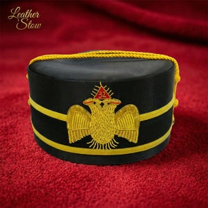 Puede incluir: Un sombrero negro con ribete dorado y un diseño de águila bordado en oro. El sombrero tiene un ribete de cuerda amarilla y un triángulo rojo con el número 32. El texto "Leather Stow" está en la esquina superior izquierda.