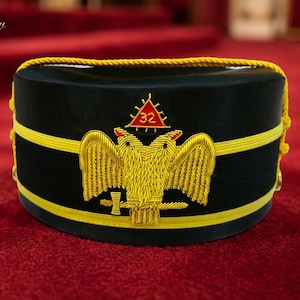 Puede incluir: Un sombrero negro con ribete dorado y un emblema de águila bicéfala bordado en oro. El sombrero tiene un triángulo rojo con el número 32. El texto "Leather Stow" está en la esquina superior izquierda.