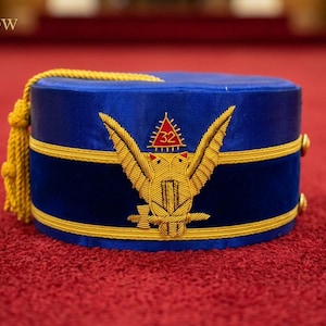 Puede incluir: Gorra de terciopelo azul con ribete dorado y un emblema bordado en oro con un pájaro y el número 32. La gorra tiene una borla dorada y el texto "Leather Stow".