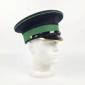 Pode incluir: Um chapéu azul marinho e verde de estilo militar com viseira preta e acabamento dourado. O chapéu é exibido em uma cabeça de manequim branca. O chapéu tem uma faixa verde ao redor da coroa.