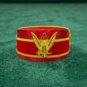 Puede incluir: Un fez rojo con bordados y adornos dorados. El sombrero presenta un emblema dorado de un águila con el número 32 y otros símbolos. El sombrero está sobre una superficie verde. El texto "Leather Stow" está en la esquina superior izquierda.
