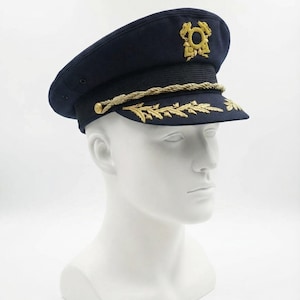 May include: A navy blue hat with gold embroidery and a gold rope trim. The hat is displayed on a white mannequin head. The hat features a gold emblem and gold leaf-like embroidery on the brim.