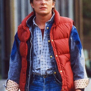 Puede incluir: Un chaleco acolchado rojo con botones dorados sobre una camisa a cuadros azul y una chaqueta vaquera. Las mangas de la chaqueta vaquera están enrolladas, revelando un forro estampado. La persona lleva vaqueros azules.