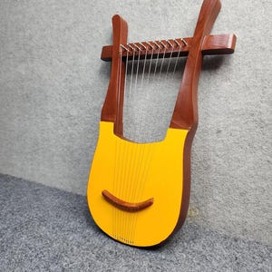 May include: A wooden lyre featuring a dark brown frame and a bright yellow soundboard. The instrument has eight thin metal strings stretched vertically across the body, secured by silver tuning pins at the top and a curved wooden bridge.
