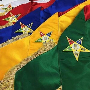 Puede incluir: Una colección de bandas de satén superpuestas en blanco, rojo, azul, amarillo y verde. Cada banda presenta un fleco de tono dorado en el borde inferior y un emblema bordado de estrella de cinco puntas con varios símbolos en la tela.