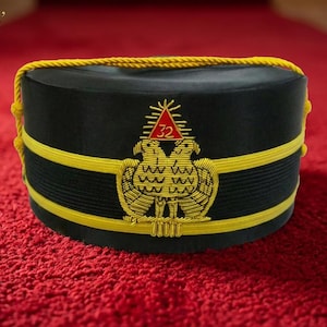 Puede incluir: Gorra negra con ribete dorado y un emblema de águila bicéfala. La gorra tiene una cuerda amarilla a lo largo del borde superior y dos bandas amarillas. Un triángulo rojo con el número 32 está encima del águila.